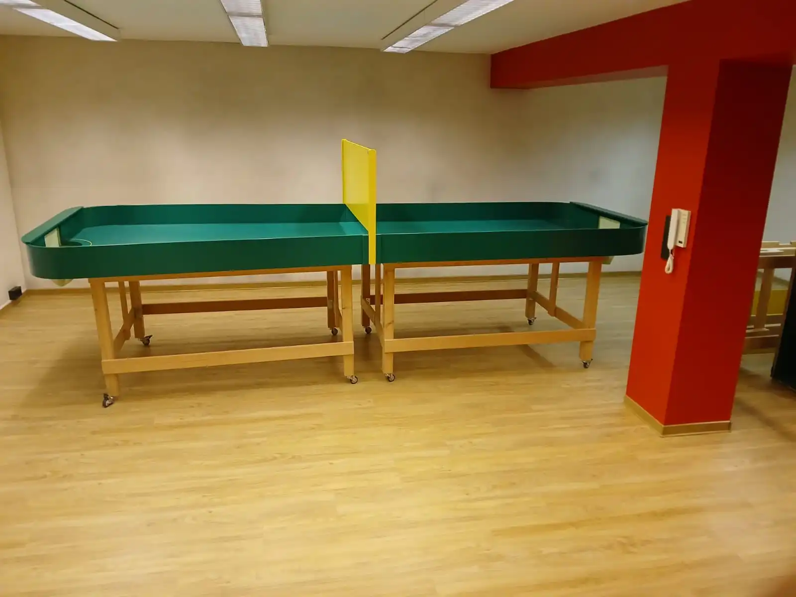 In den Vereinsräumen gibt es auch einen eigenen Sportraum mit einer Tischballplatte. Bildbeschreibung. Das Bild zeigt einen Raum mit Holzboden und einem grünen Tisch in der Mitte. Bei dem Tisch hanelt es sich um einen Showdowntisch. Der Tisch hat erhöhte Kanten und wird von Holzrahmen mit Rädern zur Beweglichkeit getragen. Rechts vom Tisch befindet sich eine rote Wand mit einer weißen Gegensprechanlage darauf. Die Decke ist mit rechteckigen Leuchten ausgestattet, und die Wände sind schlicht und hell gefärbt. Ganz rechts ist ein Teil einer weiteren Struktur oder Möbel sichtbar. Der Raum wirkt geräumig, ohne andere Gegenstände oder Möbel in unmittelbarer Nähe des Tisches.
