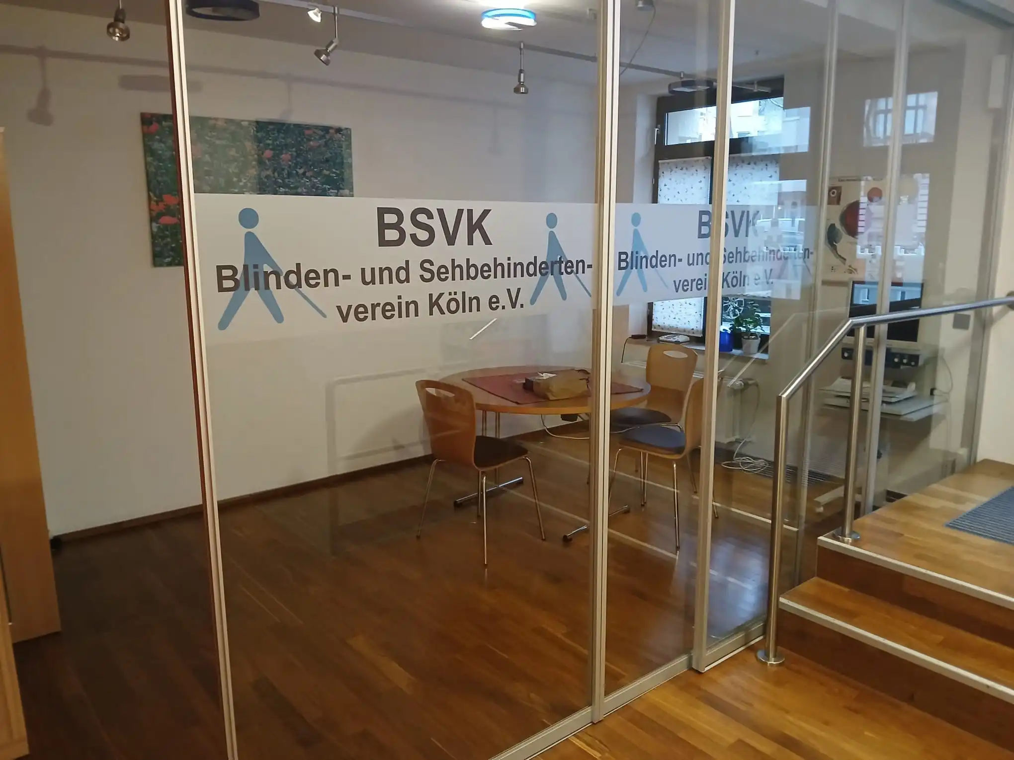 Unser getrennter Beratungsraum garantiert Vertraulichkeit während unserer Beratungszeit. Bildbeschreibung. Das Bild zeigt einen Innenraum mit einer Glastrennwand. Auf der Glaswand steht der Text BSVK Blinden- und Sehbehindertenverein Köln e.V., begleitet von einer Grafik einer Person mit einem Blindenstock. Hinter der Glastrennwand befindet sich ein kleiner Raum mit Holzboden. Im Raum steht ein runder Tisch mit vier Stühlen darum herum. Auf dem Tisch liegen einige Gegenstände, darunter eine Tasche und andere Gegenstände. Ein Gemälde ist an der Wand hinter dem Tisch angebracht, das eine Naturszene mit Blumen darstellt. An der Seite des Raums befindet sich ein Schreibtisch mit einem Computermonitor und anderen Gegenständen. Im Hintergrund ist ein Fenster mit gemusterten Vorhängen zu sehen, zusammen mit einigen Topfpflanzen, die auf der Fensterbank stehen.
