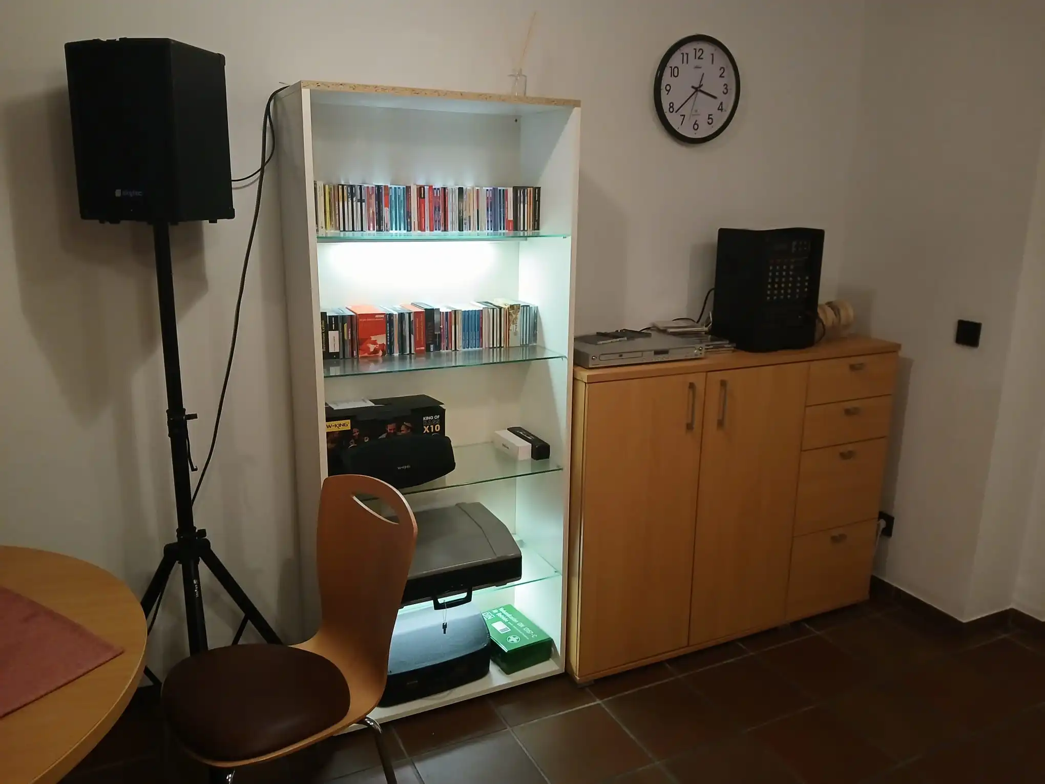 In unserem Hauptraum befindet sich auch unserer offene Hörbücherschrank. Mitglieder können sich hier jederzeit Hörbücher ausleihen und Hörbücher den Mitgliedern zur Verfügung stellen. Bildbeschreibung. Das Bild zeigt eine Ecke eines Raumes mit verschiedenen Gegenständen und Möbeln. Auf der linken Seite befindet sich ein schwarzer Lautsprecher, welcher auf einem Stativ montiert ist. Der Lautsprecher befindet sich links von einer weißen Regaleinheit. Ein Holzstuhl mit gebogener Rückenlehne und ohne Armlehnen ist vor der Regaleinheit aufgestellt. Der Stuhl steht in der Nähe eines runden Holztisches, von dem ein Teil in der unteren linken Ecke sichtbar ist. In der Mitte steht ein weißes Regal. Das Regal ist von innen beleuchtet. Das Regal enthält eine Sammlung von CDs, die in Reihen angeordnet sind. Zudem liegt hier ein schwarzer Lautsprecher bereit. Weiter unten liegen ein paar Kisten. Auf der rechten Seite befindet sich ein Holzschrank mit zwei Türen und drei Schubladen. Oben auf dem Holzschrank befinden sich ein Plattenspieler und ein schwarzes Audiosystem. Einige Kabel sind in der Nähe des Audiosystems sichtbar. Eine runde Wanduhr ist über dem Schrank montiert, die die Zeit etwa 10:10 Uhr anzeigt.