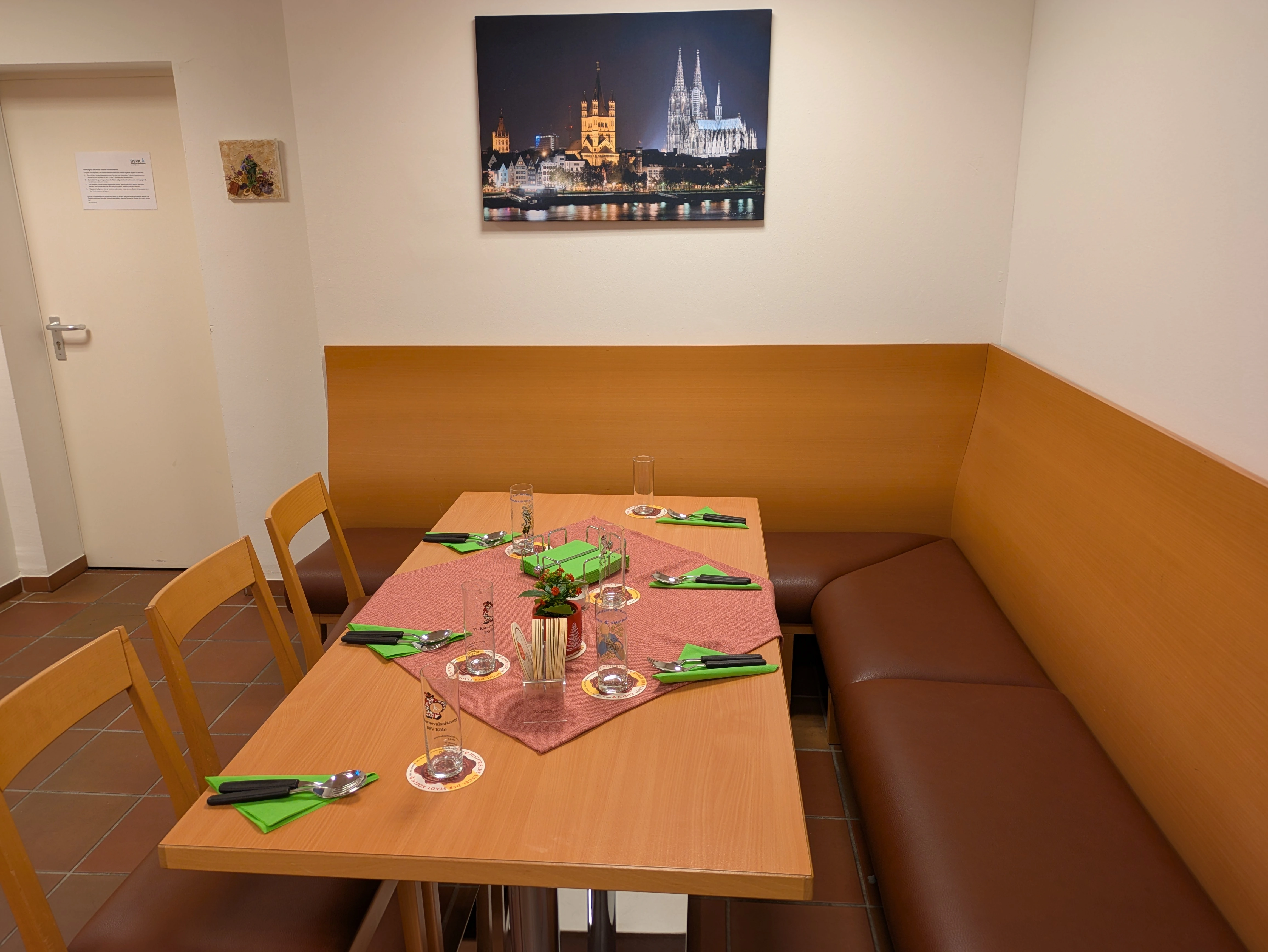 Unsere gemütliche Bankecke lädt zum entspannen ein. Bildbeschreibung. Das Bild zeigt einen kleinen Essbereich mit einem rechteckigen Holztisch, der in der Mitte steht. Der Tisch ist an drei Seiten von Sitzgelegenheiten umgeben: einer braunen gepolsterten Bank an der rechten und hinteren Wand sowie drei Holzstühlen auf der linken Seite. Der Tisch ist für sechs Personen gedeckt. Ein rosa Tischläufer ist diagonal über dem Tisch platziert, und in der Mitte befindet sich ein kleines Blumenmittelstück. Die Wände sind hell gefärbt, und ein großes gerahmtes Bild der Kölner Stadtlandschaft ist an der Wand über der Bank angebracht. Links befindet sich eine geschlossene weiße Tür mit einem Schild und eine kleine dekorative Wand daneben. Der Boden ist mit braunen quadratischen Fliesen gefliest.