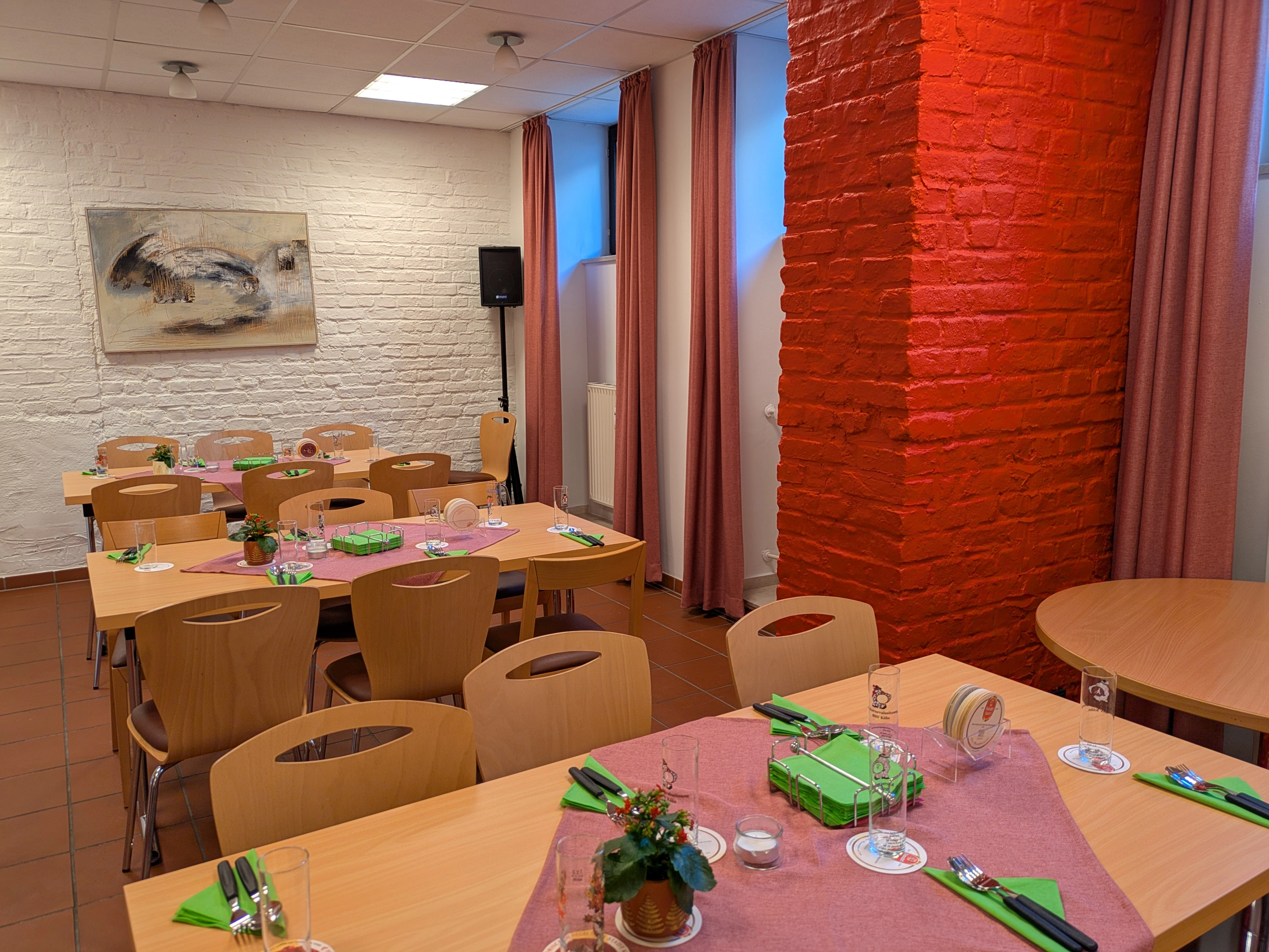 In unseren Räumlichkeiten können allerlei Veranstaltungen stattfinden. Regelmäßig treffen sich hier unsere Gruppen. Bildbeschreibung. Das Bild zeigt einen Speisesaal mit mehreren Holztischen und Stühlen, die in einem rechteckigen Raum angeordnet sind. Der Raum hat eine weiße Backsteinwand auf der linken Seite und eine rote Backsteinsäule nahe der mittleren rechten Seite. Der Boden ist rötlich-braun gefliest. Es gibt vier rechteckige Tische und einen runden Tisch. Die Tische sind mit rosa Tischläufern bedeckt, und jeder Tisch ist mit grünen Servietten, Besteck, Glasbechern und kleinen Dekorationsgegenständen gedeckt. Kleine Topfpflanzen wurden auf die Tische gestellt, zusammen mit Untersetzern und Kerzen in Glashaltern. Die linke Wand ist weiß gestrichen und zeigt ein abstraktes Gemälde. Die rote Backsteinsäule ist auffällig und teilt den Raum visuell. Die Fenster an der rechten Wand sind mit rosa Vorhängen bedeckt.