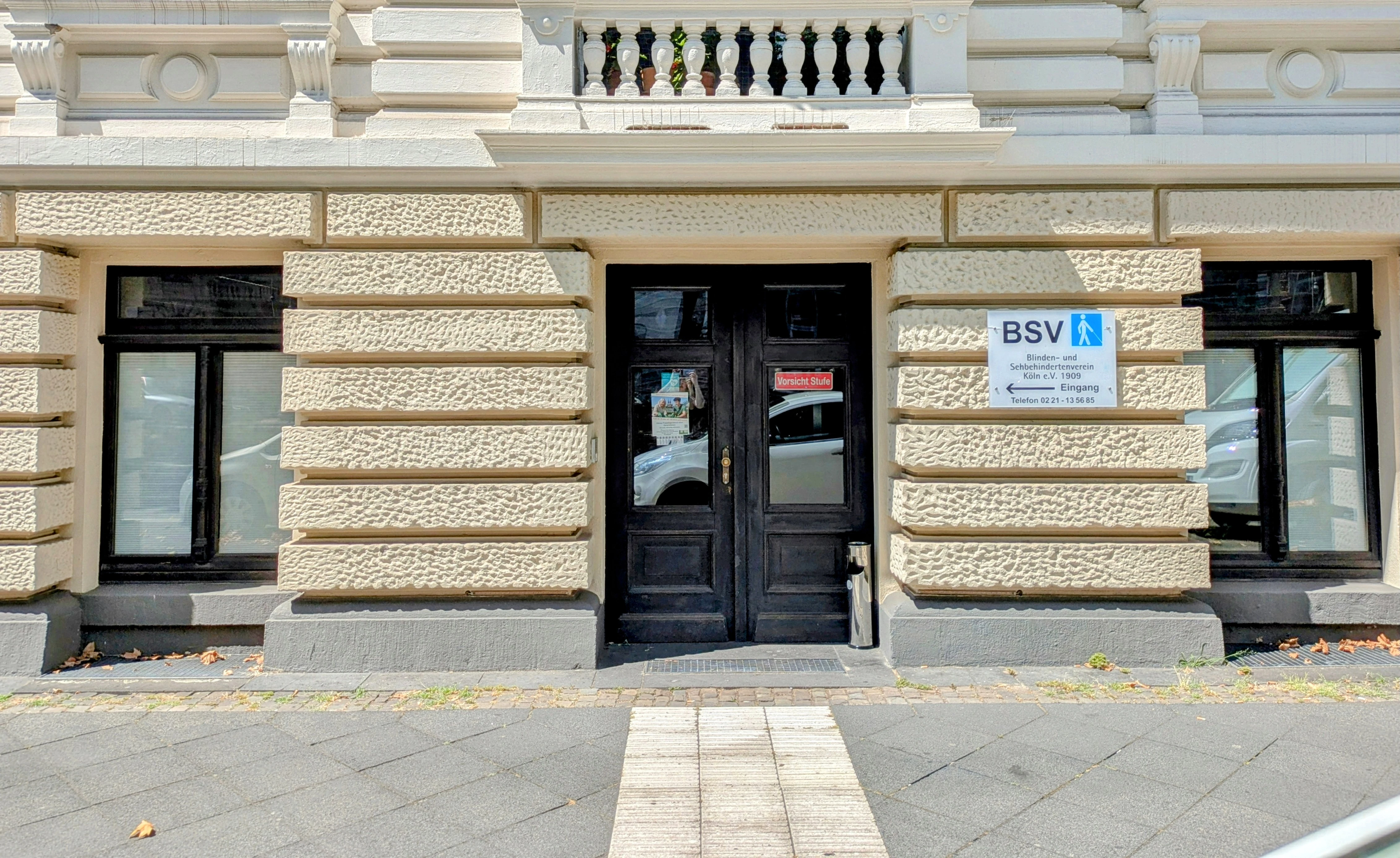 Eingang unseres Vereins am Hansaring 15. Unser Zugang hat innen zwei Treppenstufen und wir verfügen auch über einen Aufzug so das unsere Räumlichkeiten auch mit einem Rollstuhl betreten werden können. Bildbeschreibung. Das Bild zeigt die Außenfassade eines Gebäudes mit einer strukturierten Fassade mit horizontalen Rillen. Das Gebäude hat drei sichtbare Abschnitte: zwei Fenster auf jeder Seite einer zentralen Tür. Die beiden Fenster sind rechteckig und haben schwarze Rahmen. Beide Fenster sind in die strukturierte Wand eingelassen und auf gleicher Höhe gelegen. Die zentrale Tür ist schwarz und besteht aus zwei Paneelen mit Glaseinsätzen. Ein Schild mit der Aufschrift Vorsicht Stufe ist an der oberen Glasplatte der Tür angebracht. Die Tür hat einen Messinggriff auf der rechten Seite. Rechts von der Tür, an der Wand montiert, befindet sich ein rechteckiges Schild mit der Aufschrift Blinden- und Sehbehindertenverein Köln e.V. 1908, einem Pfeil Richtung Eingang und der Telefonnummer 0221 - 13 56 85. Das Schild zeigt zudem ein blaues Symbol einer Person, die mit einem Stock geht. Der Boden vor dem Gebäude besteht aus einem gefliesten Gehweg mit einem taktilen Streifen, der zur Tür führt.